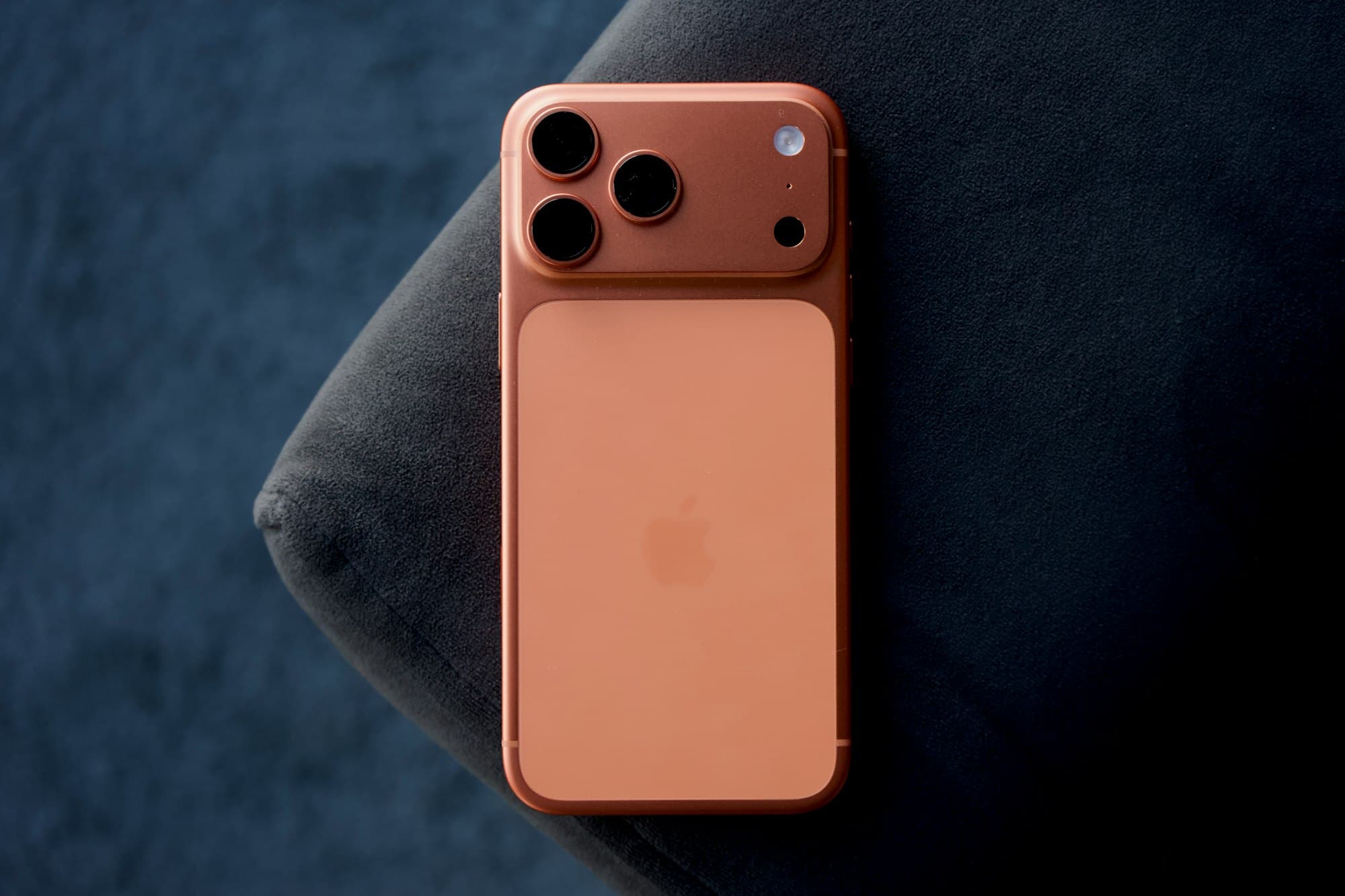 Die Rückseite eines orangefarbenen iPhone Pro mit 3 Kameras auf der Armlehne eines dunklen Sofas