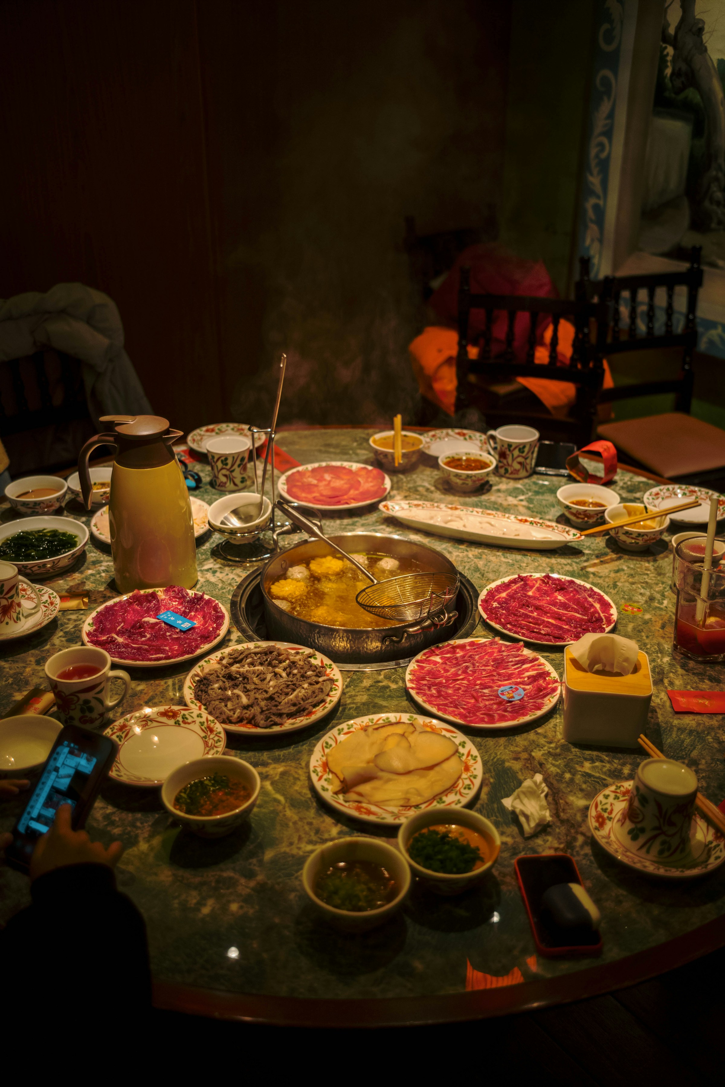 A communal hot pot table with simmering broth surrounded by plates of sliced meat, vegetables and dipping bowls